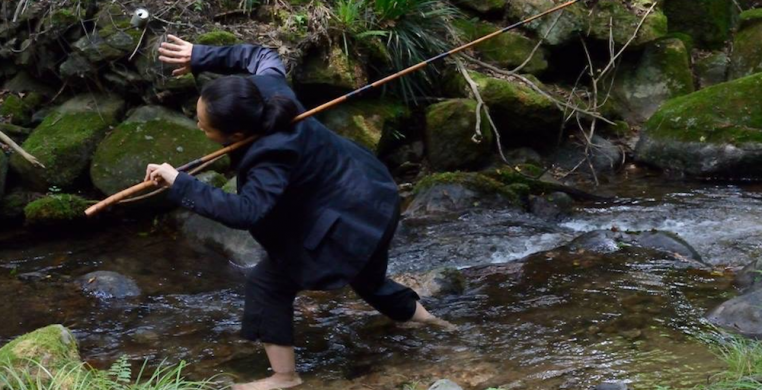 Ayako Kato by Yasunori Hangoh. A person dressed in a black suit jacket and black pants wades barefoot through a stream carrying a wooden fishing pole over one shoulder. 
