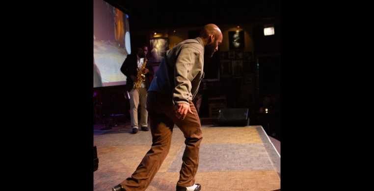 Jumaane Taylor tap dancing facing Brent Griffin Jr. playing the saxophone on stage at the Jazz Showcase.