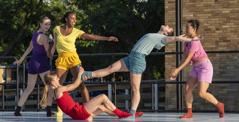 4 women dancers in brightly colored shorts and tops on an outdoor stage in front of a background of green trees. The dancer in teal is leaning far backwards, supported by a dancer in red holding her leg while two others reach for her.