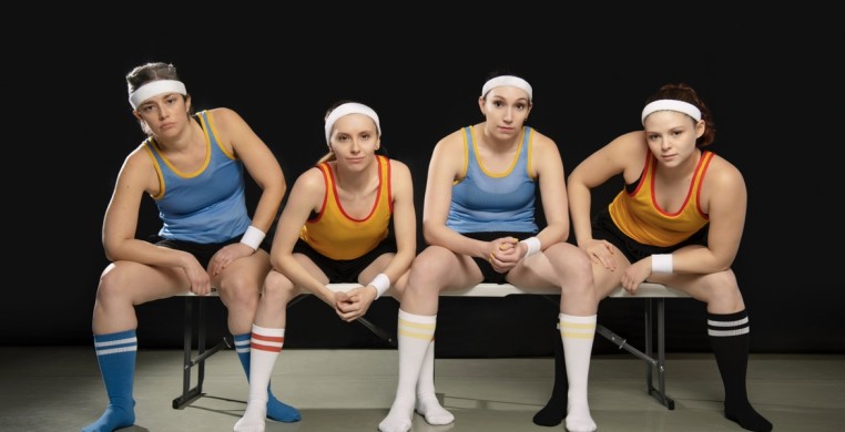 Photo by Ashley Deran. Four dancers sit on a bench in casual positions. They are wearing white headbands, sports tank jerseys that alternate blue and yellow, black pants and knee high athletic socks in blue white and black . 