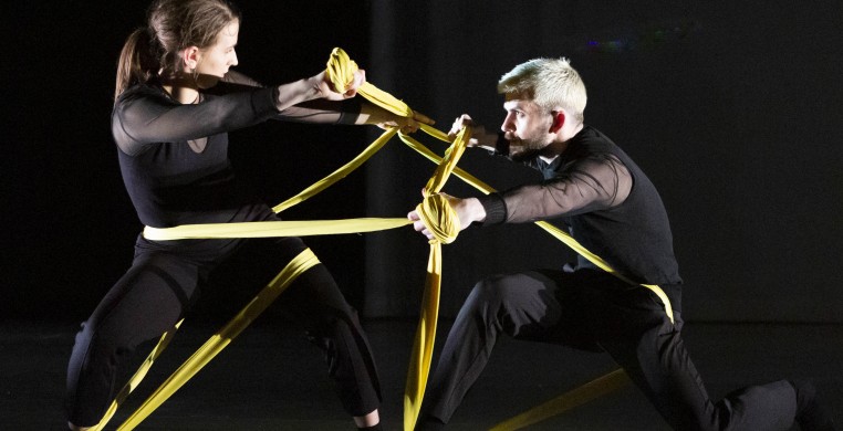 Two dancers in black face each other while intertwined with yellow fabric ribbons