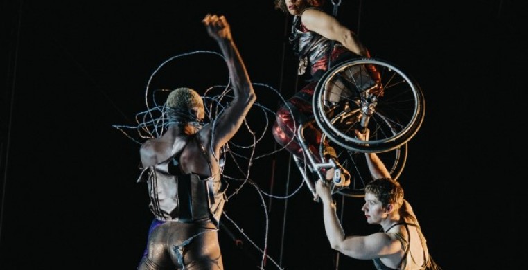 Three dancers face each other onstage. Jerron, a dark-skinned Black man with blonde hair, stands boldly facing the others, his body tense with energy. His tight pants and leather top shimmer. His fist flies overhead as silver barbe
