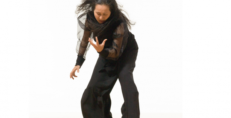 Ayako Kato dressed in all black in a white background. She is bending her knees and her gaze is on her left arm, wrist facing up.