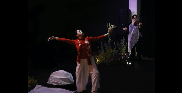 A dancer expanding her arms in the kimono fabric red shirt and white pants among Japanese flower arrangement. A violinist is playing.