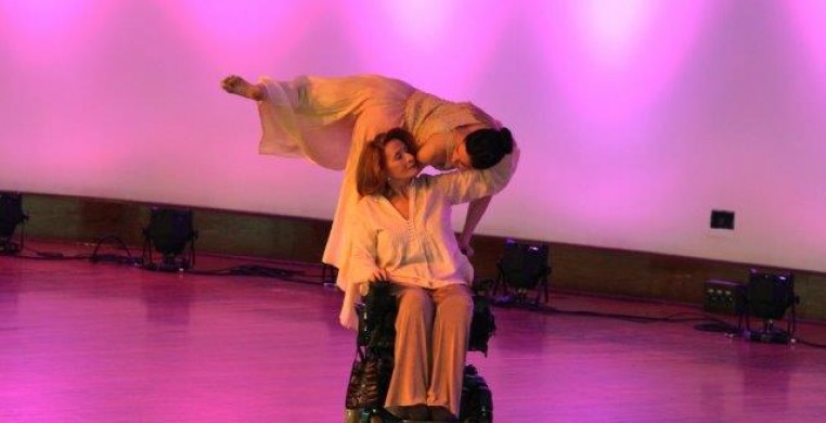 Two women in pastel, the younger climbs on the elder dancers power wheelchair. they look into each others eyes.