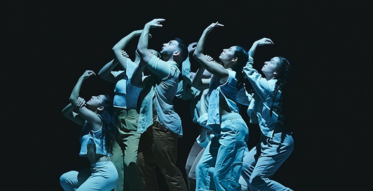 Six dancers are in a group close together in profile looking up, some crouching and some standing. All the dancers have their left arm reaching up, with their right hand clasping their left bicep.