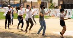 Image of seven people dancing in a line outside on a basketball court with a mural in the background