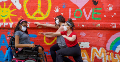 three dancers wearing white masks against a mural on a brick wall. One dancer in a power chair is on the left and two dancers lunge on the right, reaching an arm to the other dancer. Photo Credit: Justin Cooper