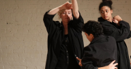 Three dancers wearing all black standing in a clump doing various gestural shapes.  