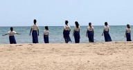 8 dancers in blue and white tunics walk from the beach into the water, against the backdrop of a blue sky. 
