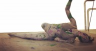 Woman painted all grey with splotches of colored paint dances on floor with arm extended