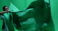 Still image of Black performer with curly hair in green costume holding the corners of a piece of green fabric.