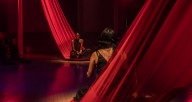 Two people sit on long red pieces of fabric facing each other in a red-lit room.